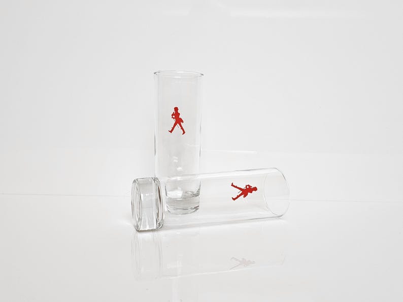 Peut inclure: Trois verres &agrave; shot transparents avec une figurine rouge d'un homme qui marche. Un verre est debout, un autre est couch&eacute; sur le c&ocirc;t&eacute;, et le troisi&egrave;me est la base d'un verre. Les verres sont sur une surface blanche.