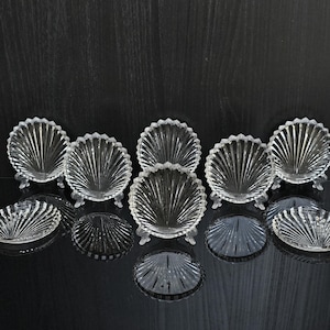 Op de afbeelding: Een verzameling heldere glazen schelpvormige schalen. De schalen hebben een geribbeld ontwerp en zijn gerangschikt op een reflecterend zwart oppervlak. Sommige schalen staan op kleine voetjes.