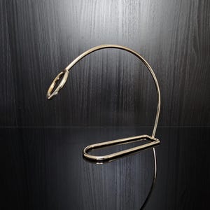 Op de afbeelding: Een goudkleurige metalen wijnfleshouder met een gebogen ontwerp. De houder heeft een lus aan de bovenkant voor de flessenhals en een rechthoekige basis. De standaard staat op een reflecterend oppervlak.