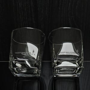 May include: Two clear, square-shaped whiskey glasses with the text "KEEP WALKING" printed on the side. The glasses are made of thick glass and have a textured design. The background is dark.