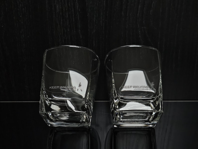 May include: Two clear, square-shaped whiskey glasses with the text "KEEP WALKING" printed on the side. The glasses are made of thick glass and have a textured design. The background is dark.