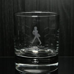 May include: A clear, lowball whiskey glass with the Johnnie Walker logo etched on the front. The logo features a silhouette of a man walking with a cane. The glass has a thick base and a rounded shape.