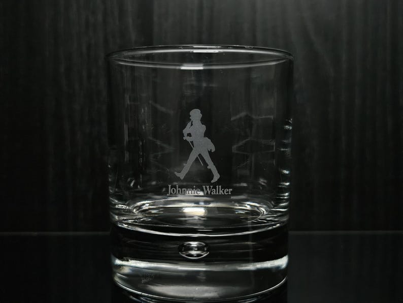 May include: A clear, lowball whiskey glass with the Johnnie Walker logo etched on the front. The logo features a silhouette of a man walking with a cane. The glass has a thick base and a rounded shape.