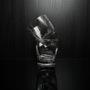 May include: Two clear, square-shaped whiskey glasses stacked on a reflective black surface. The glasses have a geometric design and are partially filled. The background is a dark wood grain.