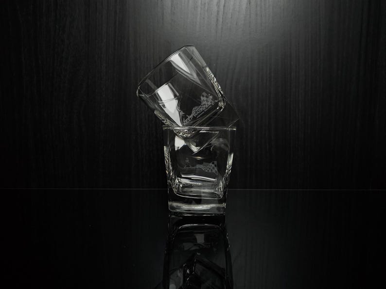 May include: Two clear, square-shaped whiskey glasses stacked on a reflective black surface. The glasses have a geometric design and are partially filled. The background is a dark wood grain.