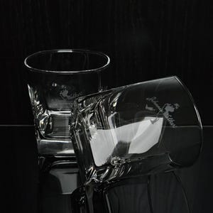 May include: Two clear, square-shaped whiskey glasses with the "Johnny Walker" logo etched on the side. One glass is upright, the other is tilted, reflecting light. The glasses are on a dark, reflective surface.