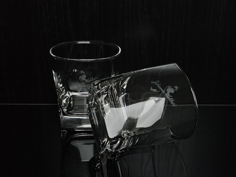 May include: Two clear, square-shaped whiskey glasses with the "Johnny Walker" logo etched on the side. One glass is upright, the other is tilted, reflecting light. The glasses are on a dark, reflective surface.