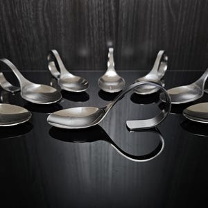 Op de afbeelding: Een verzameling roestvrijstalen lepels met een uniek ontwerp. Elke lepel heeft een lusvormige handgreep. De lepels zijn gerangschikt op een reflecterend zwart oppervlak. Ontworpen om te serveren of te eten.