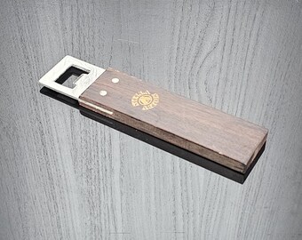 May include: A wooden bottle opener with a metal opener on one end. The wood is dark brown and has a gold logo on it.
