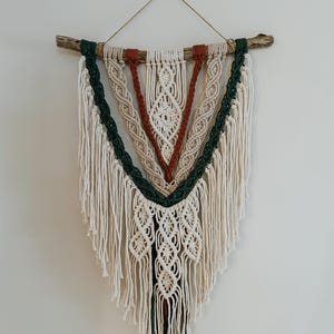 May include: A macrame wall hanging with a white, cream, green, and orange color scheme. The hanging is made with a variety of knots and has a fringe at the bottom. It is hung on a branch.