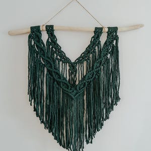 May include: A dark green macrame wall hanging with a wooden dowel at the top. The hanging features a V-shaped design with fringe details.