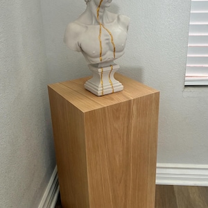May include: A white bust of a man with gold detailing, displayed on a light brown wooden plinth. The bust features gold lines. The plinth is a square, solid block of wood.