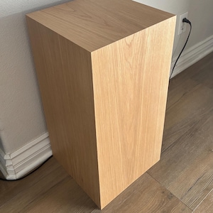 May include: A light oak wooden pedestal table with a square top and a rectangular base. The table is positioned against a white wall. A black power cord is visible. The table's dimensions are in centimetres.