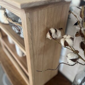 K&ouml;nnte beinhalten: Ein helles Holzregal mit drei Regalb&ouml;den. Das Regal ist aus Holz mit einer hellen Holzmaserung. Das Regal steht auf einer Holzoberfl&auml;che. Auf der rechten Seite des Bildes befindet sich ein Baumzweig mit wei&szlig;en Baumwollkugeln.