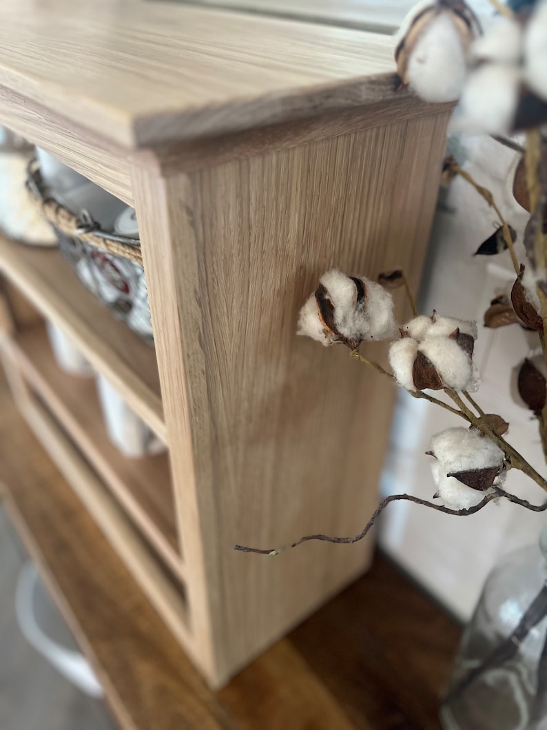 K&ouml;nnte beinhalten: Ein helles Holzregal mit drei Regalb&ouml;den. Das Regal ist aus Holz mit einer hellen Holzmaserung. Das Regal steht auf einer Holzoberfl&auml;che. Auf der rechten Seite des Bildes befindet sich ein Baumzweig mit wei&szlig;en Baumwollkugeln.