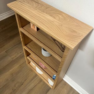 K&ouml;nnte beinhalten: Ein Wandregal aus Holz mit vier Regalbrettern. Das Regal ist aus hellem Holz mit einer nat&uuml;rlichen Oberfl&auml;che. Die Regalbretter sind leer, bis auf einen kleinen Pflanzentopf auf dem dritten Regal.
