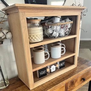 K&ouml;nnte beinhalten: Ein dreist&ouml;ckiges Holzregal mit einem durchsichtigen Glasgef&auml;&szlig;, einem Drahtkorb gef&uuml;llt mit wei&szlig;en Keramikbechern und wei&szlig;en Keramikbechern auf den Regalbrettern. Das Regal hat eine helle Holzoptik und ist mit Holzschildchen verziert, auf denen "Stay" und "Cozy" steht.