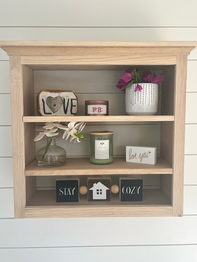 K&ouml;nnte beinhalten: Ein dreist&ouml;ckiges Holzregal mit verschiedenen Dekoartikeln, darunter ein herzf&ouml;rmiges Holzschild mit der Aufschrift "LOVE", eine kleine Kerze, ein kleines wei&szlig;es Schild mit der Aufschrift "love you" und drei schwarze Bl&ouml;cke mit den Worten "STAY", "HOME" und "COZY" darauf.