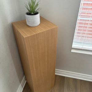 May include: A light brown wooden pedestal with a faux succulent plant in a white and grey striped pot. The pedestal is rectangular and placed in a corner against a grey wall and light brown wood floor.