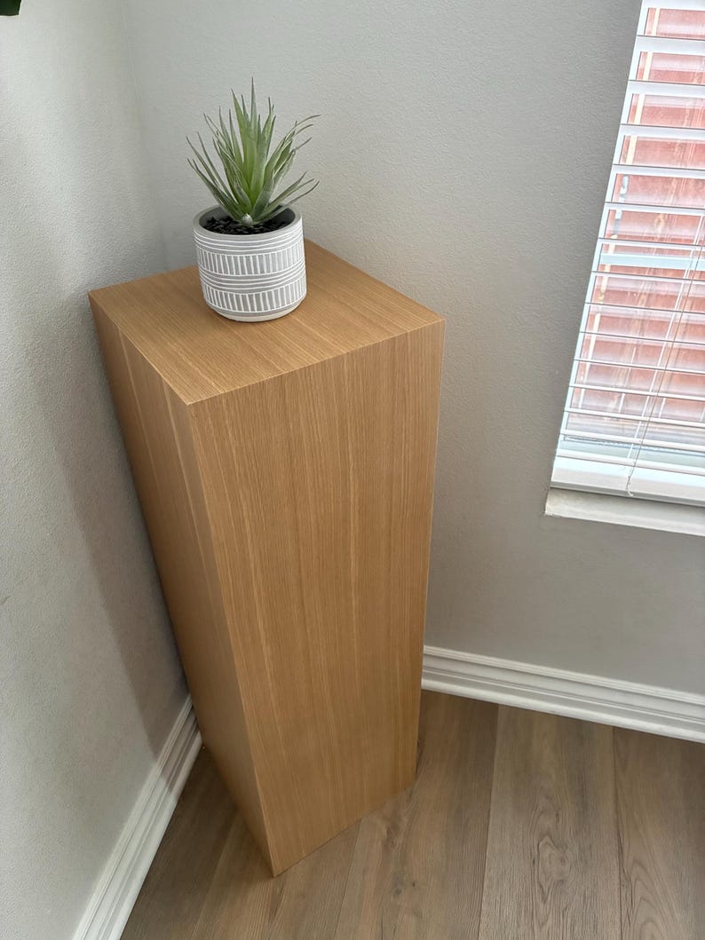 May include: A light brown wooden pedestal with a faux succulent plant in a white and grey striped pot. The pedestal is rectangular and placed in a corner against a grey wall and light brown wood floor.