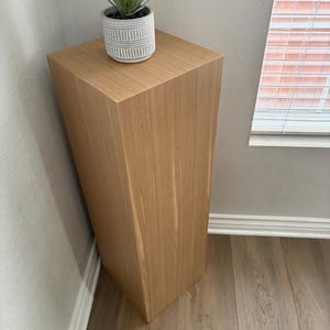 Op de afbeelding: Een lichtbruine houten sokkeltafel met een vierkant blad en een taps toelopend ontwerp. Een kleine, grijs en wit gestreepte plantenbak met een groene vetplant staat op de tafel. De tafel staat in een hoek van een kamer.