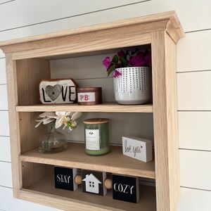 K&ouml;nnte beinhalten: Ein Wandregal aus Holz mit drei Ablagen. In der obersten Ablage befindet sich ein rotes herzf&ouml;rmiges Schild mit der Aufschrift &bdquo;LOVE&ldquo;, eine kleine rosa und rote Dose und ein wei&szlig;er Topf mit lila Blumen. In der mittleren Ablage steht eine wei&szlig;e Vase mit wei&szlig;en Blumen, eine gr&uuml;ne Kerze und ein wei&szlig;es Schild mit der Aufschrift &bdquo;love you&ldquo;. In der untersten Ablage befinden sich drei schwarze Schilder mit der Aufschrift &bdquo;STAY&ldquo;, &bdquo;COZY&ldquo; und ein Haus mit einer wei&szlig;en T&uuml;r.