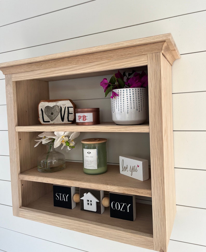 K&ouml;nnte beinhalten: Ein Wandregal aus Holz mit drei Ablagen. In der obersten Ablage befindet sich ein rotes herzf&ouml;rmiges Schild mit der Aufschrift &bdquo;LOVE&ldquo;, eine kleine rosa und rote Dose und ein wei&szlig;er Topf mit lila Blumen. In der mittleren Ablage steht eine wei&szlig;e Vase mit wei&szlig;en Blumen, eine gr&uuml;ne Kerze und ein wei&szlig;es Schild mit der Aufschrift &bdquo;love you&ldquo;. In der untersten Ablage befinden sich drei schwarze Schilder mit der Aufschrift &bdquo;STAY&ldquo;, &bdquo;COZY&ldquo; und ein Haus mit einer wei&szlig;en T&uuml;r.