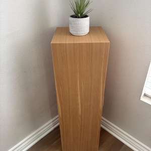 May include: A light brown wooden pedestal plant stand with a square top and a faux succulent plant in a white and grey striped pot. The stand is placed in a corner against a light grey wall.
