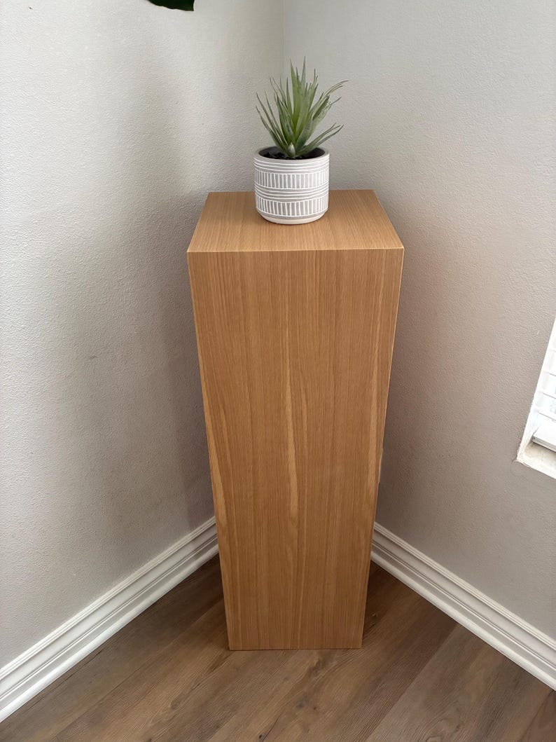 May include: A light brown wooden pedestal plant stand with a square top and a faux succulent plant in a white and grey striped pot. The stand is placed in a corner against a light grey wall.