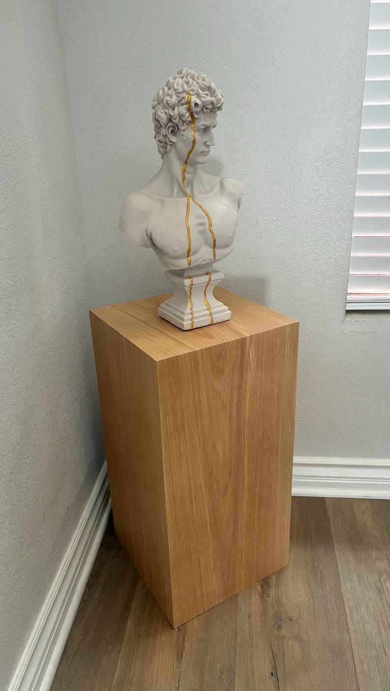May include: A white bust of a man with gold detailing, displayed on a light brown wooden plinth. The bust features gold lines. The plinth is a square, solid block of wood.