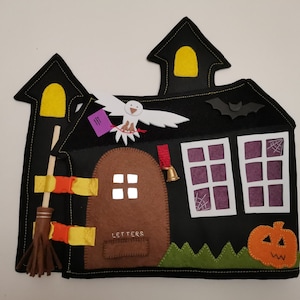 Puede incluir: Un juego de casa de Halloween de fieltro con una puerta marrón, dos ventanas, un murciélago, un búho blanco, una escoba y una calabaza. La casa tiene una luz amarilla en la ventana superior y las palabras "LETTERS" en la puerta.