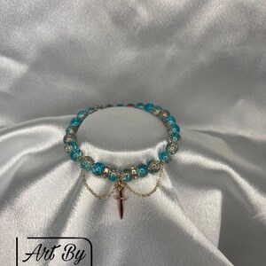 May include: A bracelet with blue and clear beads, gold-coloured metal accents, and a small dagger charm. The bracelet is presented on white, silky fabric. The text "Art By Sarah" is visible in the lower left corner.