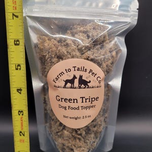 May include: A transparent plastic pouch filled with green tripe dog food topper. The pouch has a label with the text "Farm to Tails Pet Co" and "Green Tripe Dog Food Topper". The net weight is 71 grams. A yellow tape measure is next to the pouch.