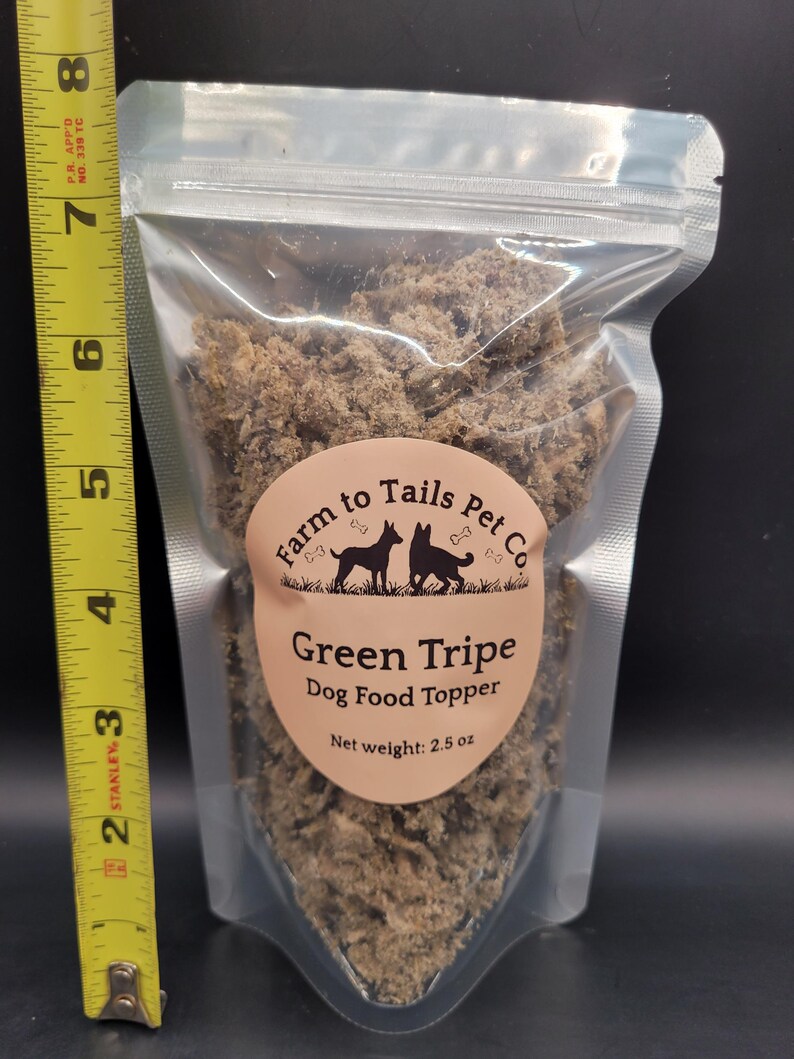 May include: A transparent plastic pouch filled with green tripe dog food topper. The pouch has a label with the text "Farm to Tails Pet Co" and "Green Tripe Dog Food Topper". The net weight is 71 grams. A yellow tape measure is next to the pouch.