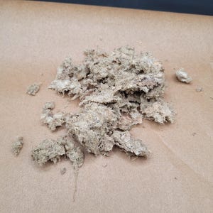 May include: A collection of light brown, fibrous material, possibly dried plant matter, is arranged on a sheet of tan paper. The material has a rough, textured appearance with irregular shapes and varying shades of brown.