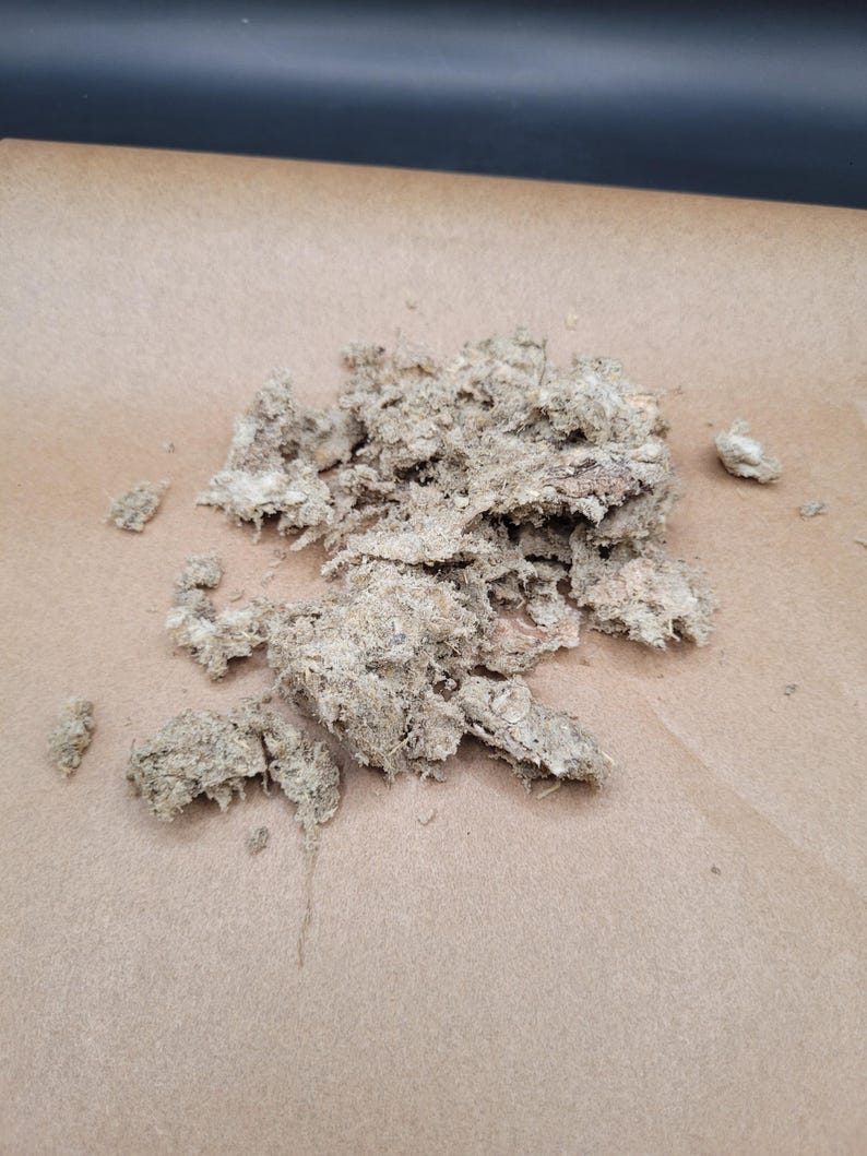 May include: A collection of light brown, fibrous material, possibly dried plant matter, is arranged on a sheet of tan paper. The material has a rough, textured appearance with irregular shapes and varying shades of brown.