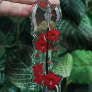 May include: Clear acrylic keychain with a silver-colored chain. The keychain features a design of a gray axe with red flowers and petals. The background is a blurred image of green leaves.