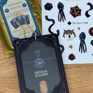 May include: A collection of tabletop gaming accessories, including a black and gold card holder with "CRITICAL SUCCESS" text, a "CHARISMA CHECK" card, and a sheet of stickers with dice and other fantasy-themed graphics. The items are on a light-colored surface.