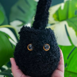 Puede incluir: Un peluche de gato negro hecho a mano con grandes ojos expresivos de color naranja y negro. El peluche tiene una textura esponjosa y una pequeña cola vertical. El fondo presenta follaje verde y blanco.