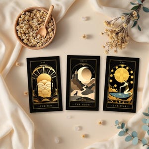 Könnte beinhalten: Drei rechteckige Tarotkarten mit schwarzem Hintergrund und Goldfolien-Illustrationen. Die Karten sind mit XIX The Sun, XVIII The Moon und XVII The Star beschriftet und zeigen Himmelsmotive, einen Wolf und eine Figur, die Wasser in ein Becken gießt.