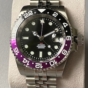 May include: A silver watch with a black face and a purple bezel. The watch has a date window at the 3 o'clock position and a GMT function. The text on the watch face reads "GMT AUTOMATIC WATER RESISTANT 20BAR/200M".
