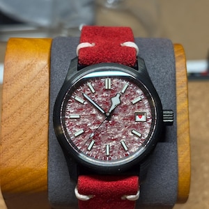 May include: A black wristwatch with a red textured face and a red suede band. The watch has white hour markers and hands, and a small red heart detail. The watch is displayed on a wooden watch stand.