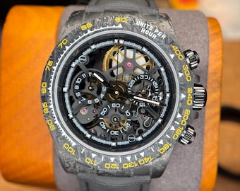 May include: A skeletonized chronograph wristwatch with a dark gray marbled carbon fiber case and bezel. The bezel features yellow tachymeter markings and the text UNITS PER HOUR. It has a black dial with visible gears, sub-dials, and a gray strap.