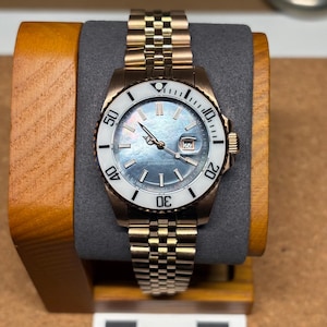 May include: A gold-coloured wristwatch with a white bezel and a blue-grey face. The watch has a date display at the 3 o'clock position and a gold-coloured strap. The watch is displayed on a wooden stand with a grey cushion.