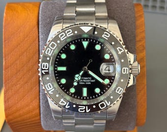 May include: A stainless steel automatic wristwatch with a black dial, luminous hour markers, and a rotating bezel featuring a 24-hour scale. The watch includes a date window at the three o'clock position and is marked as water resistant to 330 feet.