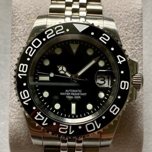 May include: A black and silver men's wristwatch with a black face, silver hands, and a silver band. The watch has a date window at the 3 o'clock position and a rotating bezel with white numbers. The watch face also has the text "Automatic Water Resistant 100m: 330ft".