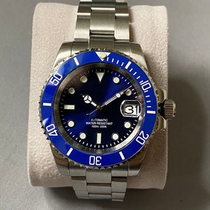 Könnte beinhalten: Eine silberne Uhr mit blauer Lünette und blauem Zifferblatt. Die Uhr hat ein Datumsfenster bei 3 Uhr und den Text "AUTOMATIC WATER RESISTANT 100m:330ft" auf dem Zifferblatt.