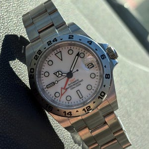 May include: A stainless steel wristwatch with a white dial, circular hour markers, and a fixed 24-hour bezel. The dial features a red GMT hand, a date window at the three o'clock position, and text reading GMT AUTOMATIC WATER RESISTANT 100m:330ft.