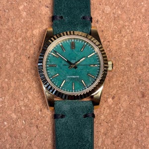 May include: A gold-toned wristwatch with a teal face and a green leather band. The watch face has gold markers and hands. The band is a deep green color with black stitching. The word "AUTOMATIC" is printed on the watch face.