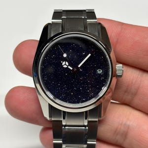 May include: A stainless steel wristwatch with a dark blue face speckled with glitter, resembling a starry night sky. The watch has silver hands and a date display at the 3 o'clock position. The watch band is also stainless steel.